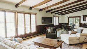 Ein geräumiges Wohnzimmer mit hohen Decken, Holzbalken, einem großen Fenster, einem Fernseher und einer Essecke mit Stühlen und einem Tisch.