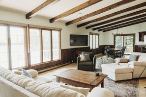 Una sala de estar amplia con techo alto, vigas de madera, un ventanal grande, un televisor y un comedor con sillas y mesa.
