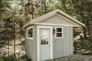 A small wooden shed with a door and two windows, surrounded by trees.