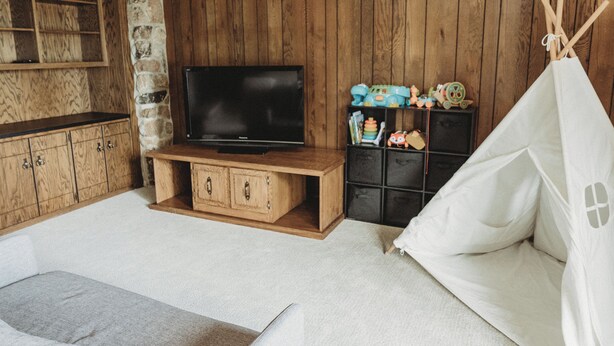 Ein Zimmer mit Fernseher auf einem Holzbalken, einer Steinwand, Holzschränken, einem Bücherregal mit Spielzeug und einer weißen, zeltartigen Konstruktion.