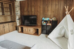 A room with a television on a wooden stand, a stone wall, wooden cabinets, a bookshelf with toys, and a white tent-like structure.
