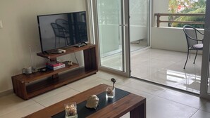 Un salon avec une table basse en bois, un téléviseur à écran plat posé sur un meuble en bois et une porte-fenêtre coulissante donnant sur un balcon équipé d’une chaise.