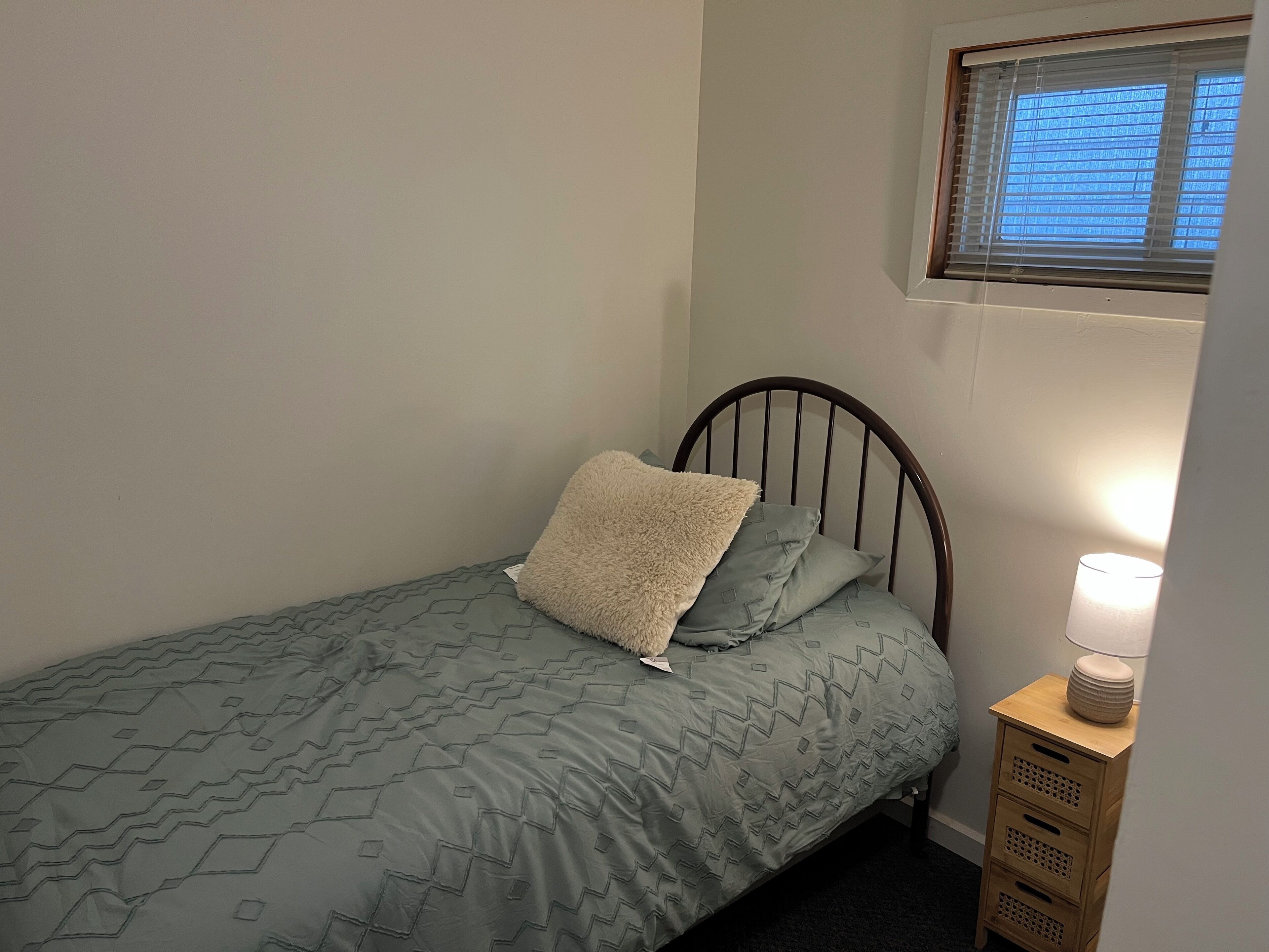 Une petite chambre avec un lit simple, recouverte d’une courtepointe grise à motifs, agrémentée d’un oreiller moelleux et d’une lampe de chevet posée sur une table de nuit en bois.