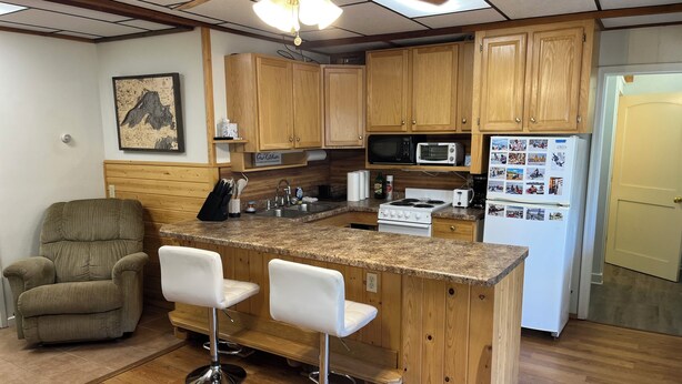Cocina con armarios de madera, microondas, estufa y refrigerador. Hay dos taburetes blancos y una silla de madera.