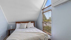 A bedroom with a sloped ceiling, a bed with white bedding, a bedside table with a lamp, and a window with blinds. - Hook Wine and Sinker (Gympie)