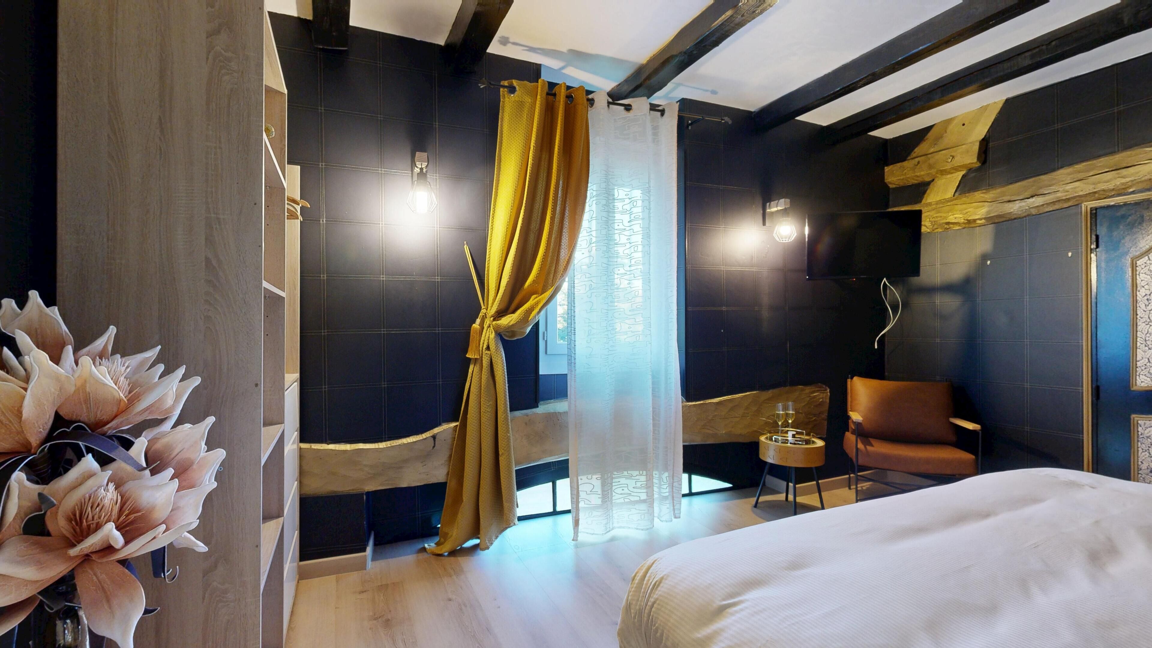 Una habitación de hotel moderna con una cama grande, una mesita con un vaso, una silla, una planta decorativa y vistas al exterior a través de una ventana con cortinas.
