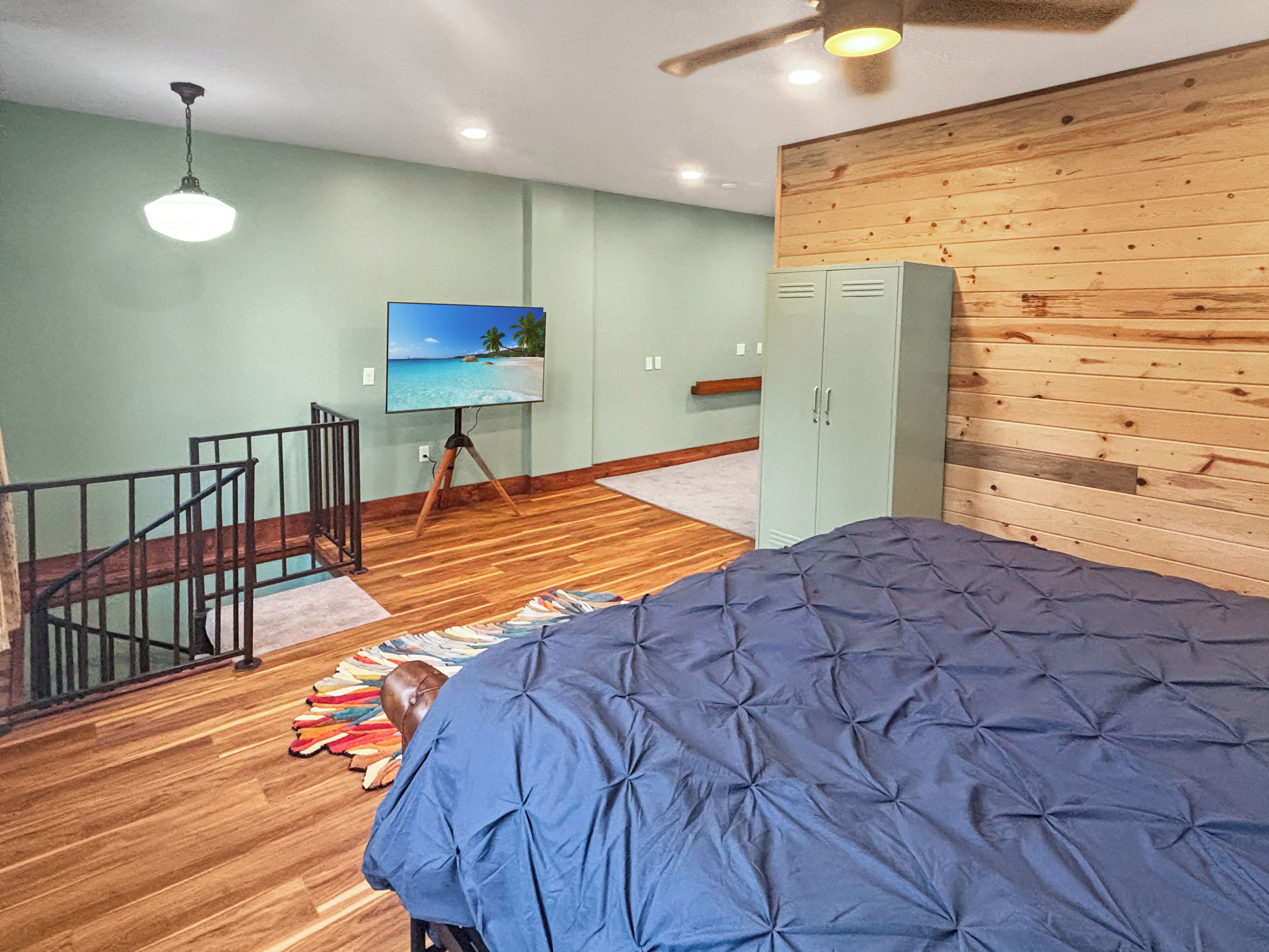 A bedroom with a large bed, a wooden wardrobe, a television on a tripod, and a staircase with a railing.