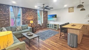 Un salon moderne avec un mur en briques, un canapé, une table basse, un téléviseur, un coin repas et un ventilateur de plafond.