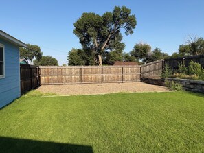 A fenced backyard with a grassy area, a wooden deck, and a large tree. - Little Blue: A Cozy 3-bedroom, 1-bath home. (Lamar)