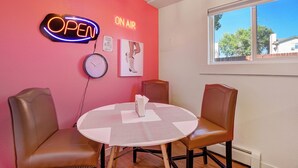 Ein Zimmer mit einer rosafarbenen Wand, einem neonbeschrifteten 'OPEN'-Schild, einer Uhr und einem Fenster mit Blick auf Bäume und ein Haus.