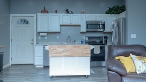 Une cuisine moderne avec des meubles blancs, un îlot central en bois, des appareils électroménagers en acier inoxydable et un canapé en cuir marron agrémenté d’un coussin jaune.