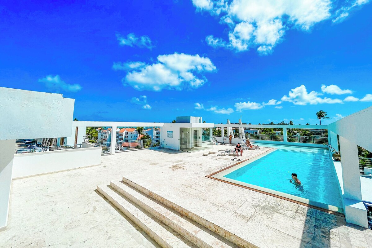 A rooftop pool area with clear blue sky and clouds, surrounded by modern buildings.