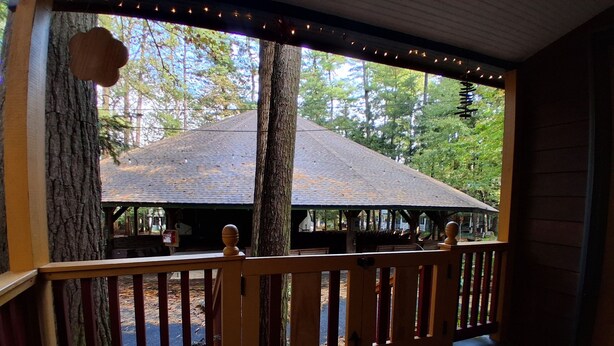 A wooden deck with a railing, a view of a building with a sloped roof, and trees in the background. - Delightful cottage in charming Mount Gretna in an amazing location! (Mount Gretna)