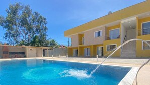 A swimming pool with a clear blue water feature, a yellow building with multiple windows, and a staircase leading to an upper level. - Condomínio Tranquilo 350m da Praia (Porto Seguro)