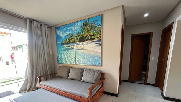 A room with a bed, a large window with curtains, a painting of a beach scene, and a door leading to another room. - Condomínio Tranquilo 350m da Praia (Porto Seguro)