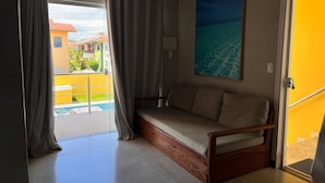 A room with a sofa, a painting, a door, and a view of a pool and buildings. - Condomínio Tranquilo 350m da Praia (Porto Seguro)