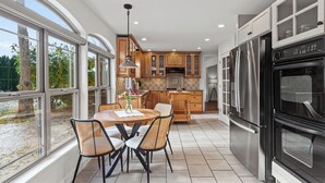 A modern kitchen with large windows, a dining area with wicker chairs, and stainless steel appliances.