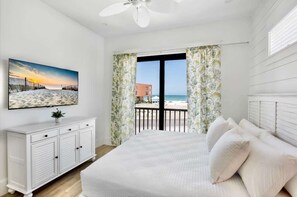 Een slaapkamer met een groot bed, een plafondventilator, een witte dressoir en uitzicht op het strand en de oceaan door het raam.