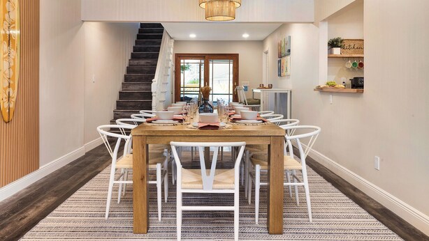 Un espace repas avec une table en bois et des chaises blanches, un escalier et une cuisine visible à travers une porte ouverte.