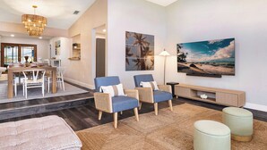 Un vaste espace de vie comprenant une table à manger, deux fauteuils bleus, des poufs, une grande peinture représentant une plage et une console en bois.
