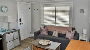 Una sala de estar con un sofá gris, una mesa de centro de madera, una puerta blanca y un ventanal con persianas.