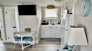 Una cocina compacta con armarios blancos, una mesa de comedor con sillas, un televisor montado sobre la encimera y un reloj en la pared.