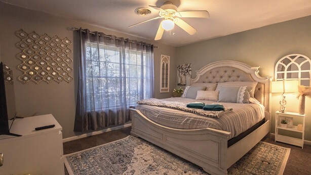 Quarto com cama branca, ventilador de teto, janela com cortinas, decoração na parede e mesa de cabeceira com abajur.
