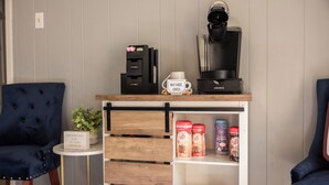 Um quarto aconchegante com um espaço para café, uma cadeira azul, uma mesinha com uma planta e uma estante com produtos de café.