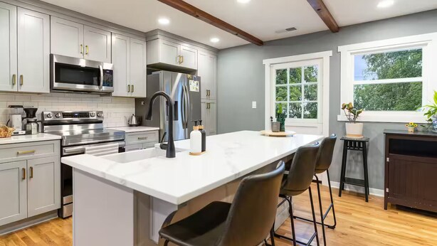 Una cocina moderna con gabinetes grises, electrodomésticos de acero inoxidable, una isla grande con taburetes de bar y una ventana con vista a árboles.
