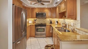 A modern kitchen with wooden cabinets, stainless steel appliances, granite countertops, and a ceiling fan. - Gorgeous 17th fl sunsets @ South Seas 2BR/2BA NEW mattresses updated furnishings (Marco Island)