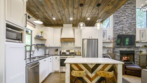 A modern kitchen with white cabinets, a central island with bar stools, a stone fireplace, and a TV mounted above it.