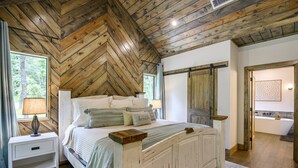 A bedroom with a herringbone wooden ceiling, a bed with pillows, a nightstand, and a view of a bathroom with a bathtub.