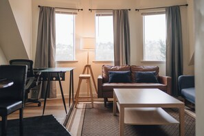 Un salon moderne avec un canapé, une table basse, un bureau et une lampe. Il y a deux fenêtres avec des rideaux.
