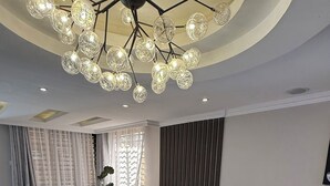 Ein modernes Wohnzimmer mit einem Ecksofa, einer Chandelüster und einem großen Fenster mit durchsichtigen Vorhängen.