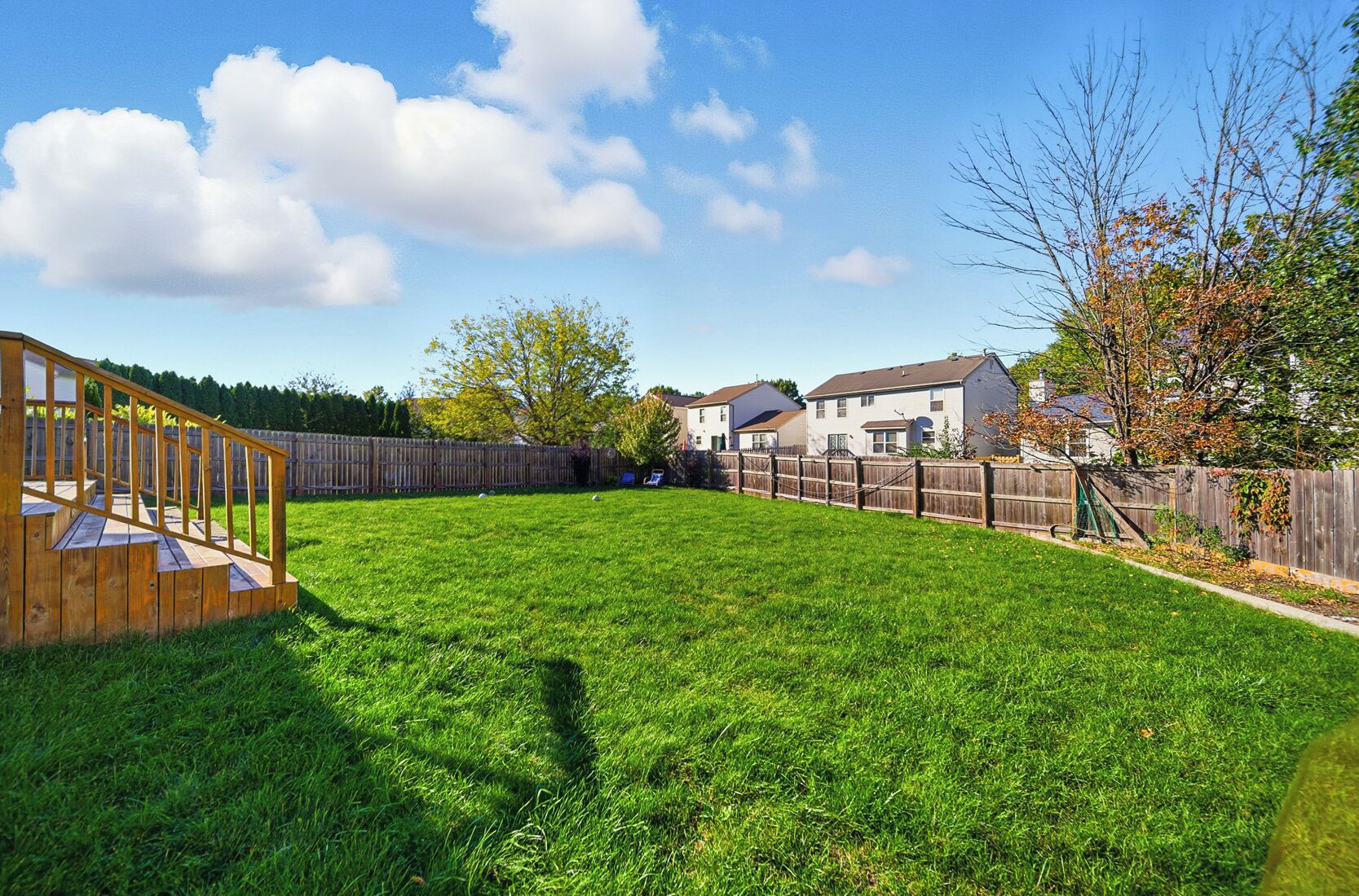 Een achtertuin met een houten terras, een gazon en een houten omheining, met huizen op de achtergrond.