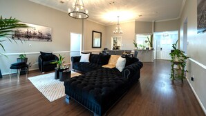 Un salon spacieux avec un grand canapé d’angle noir, un tapis blanc et plusieurs plantes en pot.