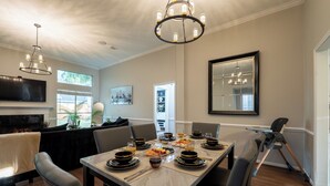 Un coin repas avec une table en marbre dressée pour six personnes, entourée de chaises. Un lustre est suspendu au plafond, un téléviseur à écran plat est fixé au mur et une cheminée est présente.