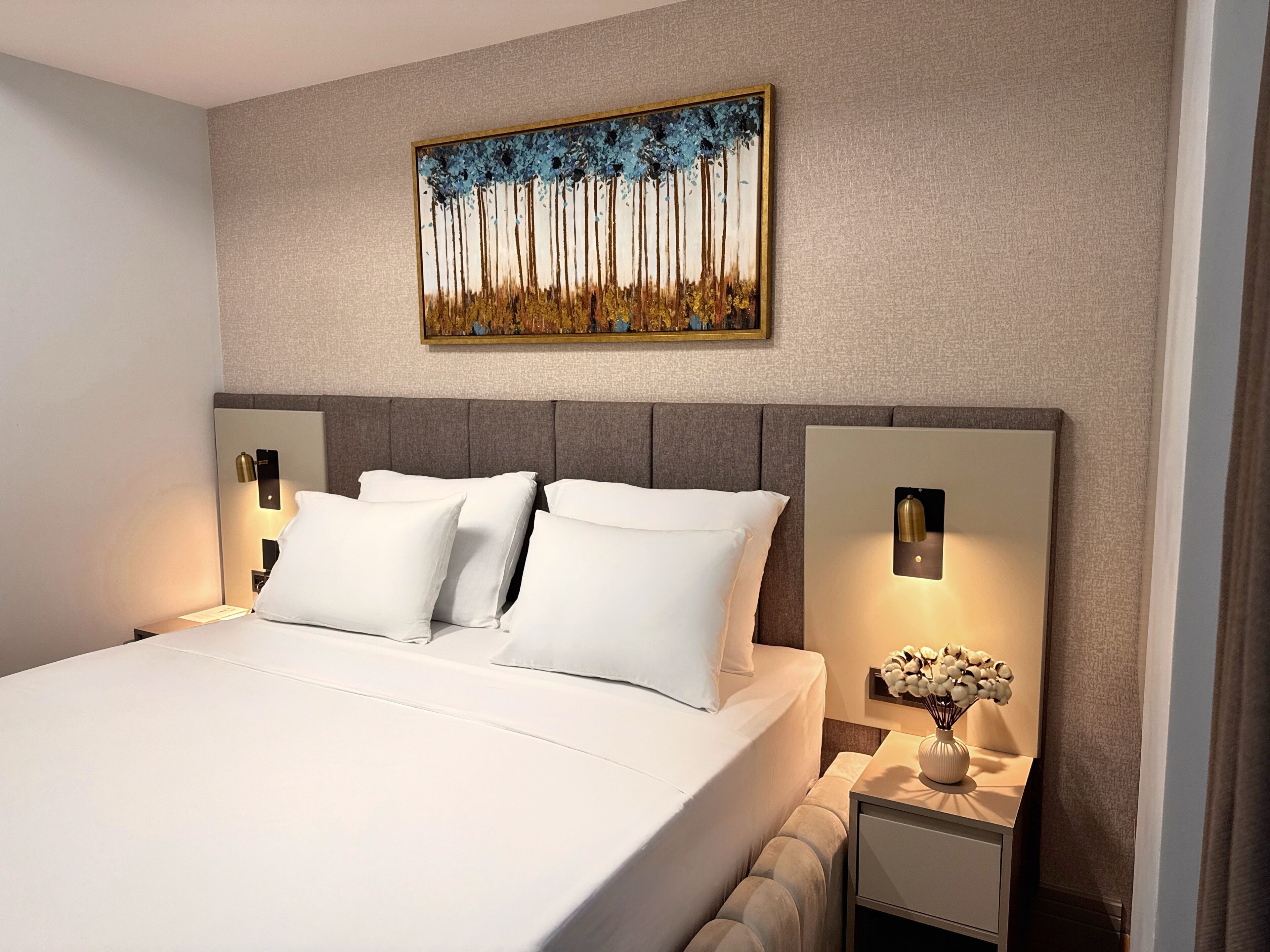 A modern hotel room with a large bed, two bedside tables with lamps, a wall-mounted painting, and a nightstand with a vase of flowers.