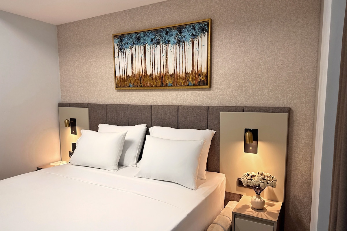 A modern hotel room with a large bed, two bedside tables with lamps, a wall-mounted painting, and a nightstand with a vase of flowers.