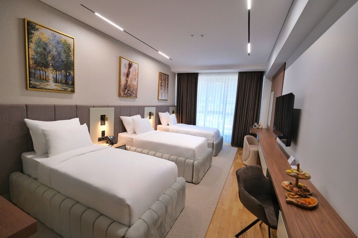 A hotel room with two beds, a desk, a chair, a TV, and artwork on the walls.