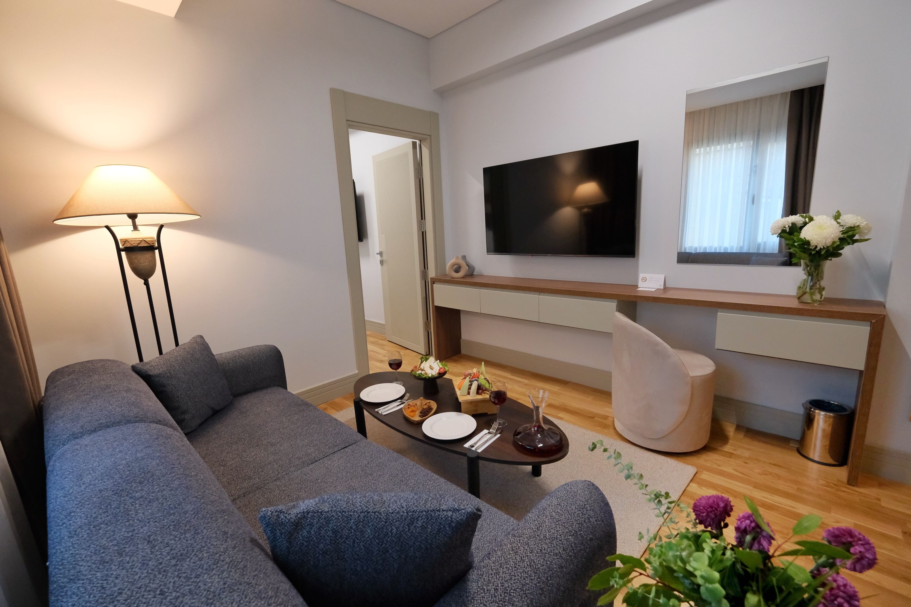 Una sala de estar moderna con un sofá gris, una mesa de centro con platos y cubiertos, una lámpara, un televisor y una ventana con un jarrón de flores.
