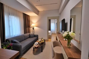 A modern hotel room with a grey sofa, a wooden coffee table with fruit, a wooden side table with flowers, a wooden console table, and a large mirror. - Park Dedeman Van (Van)