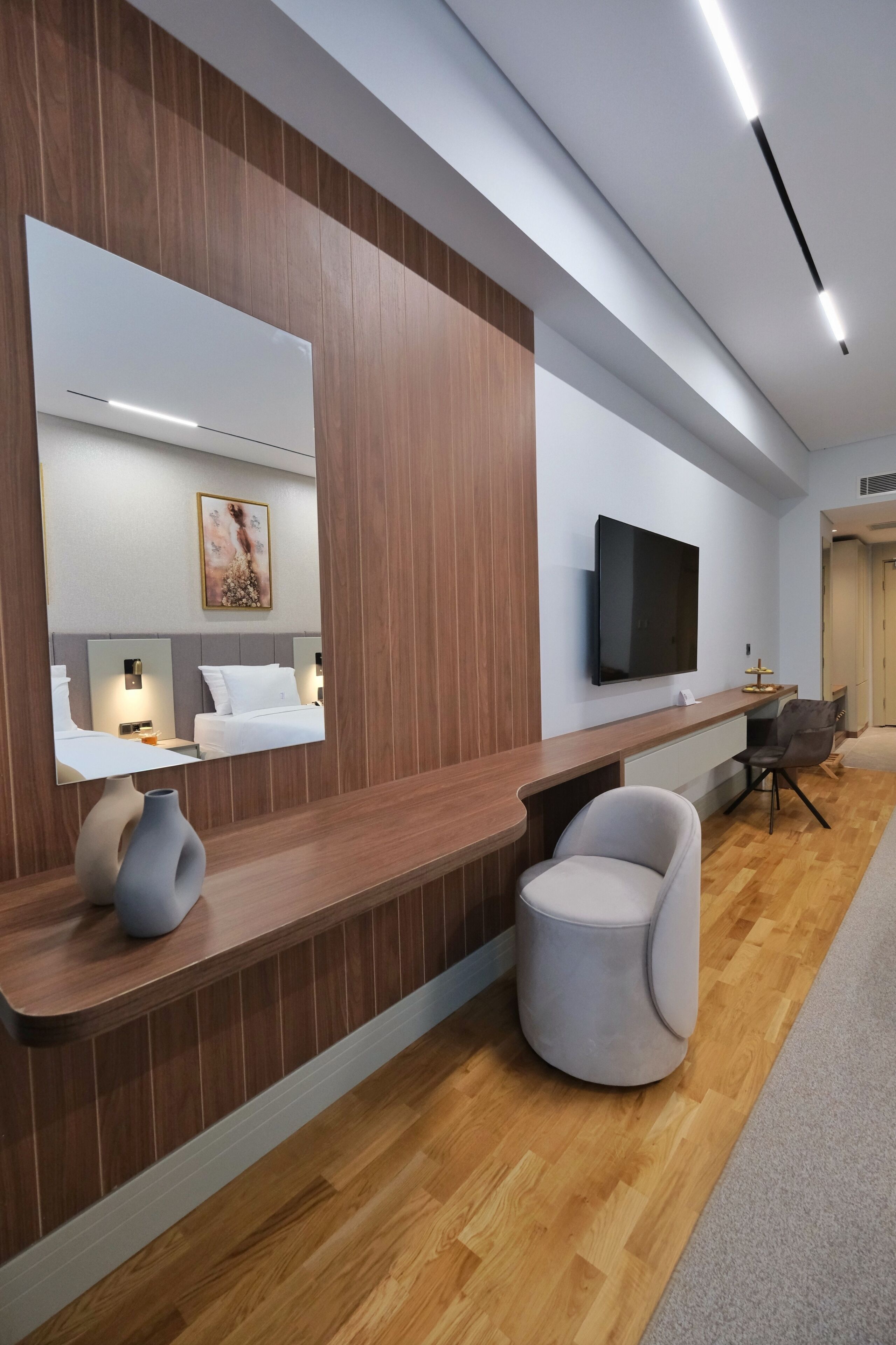 A modern hotel reception area with a wooden reception desk, a large mirror, a flat-screen TV, and a round seating area.