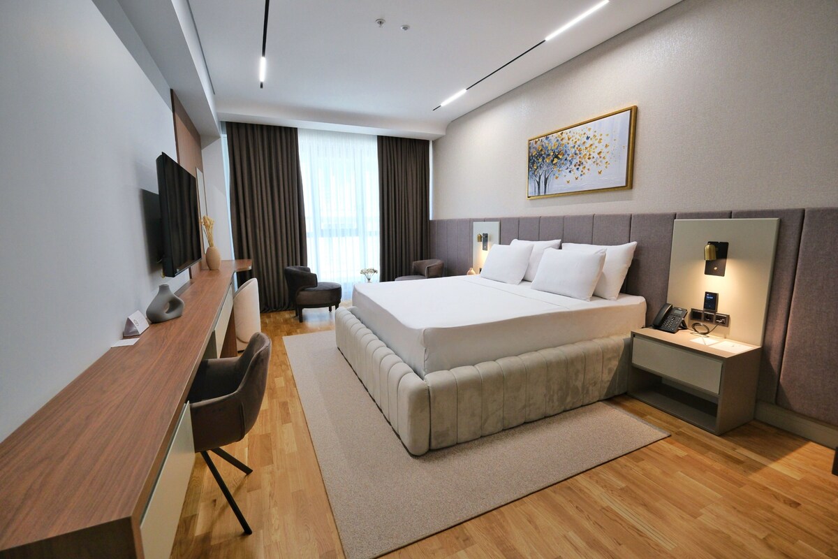 A modern hotel room with a large bed, a desk, a chair, a TV, and a bedside table with a lamp.