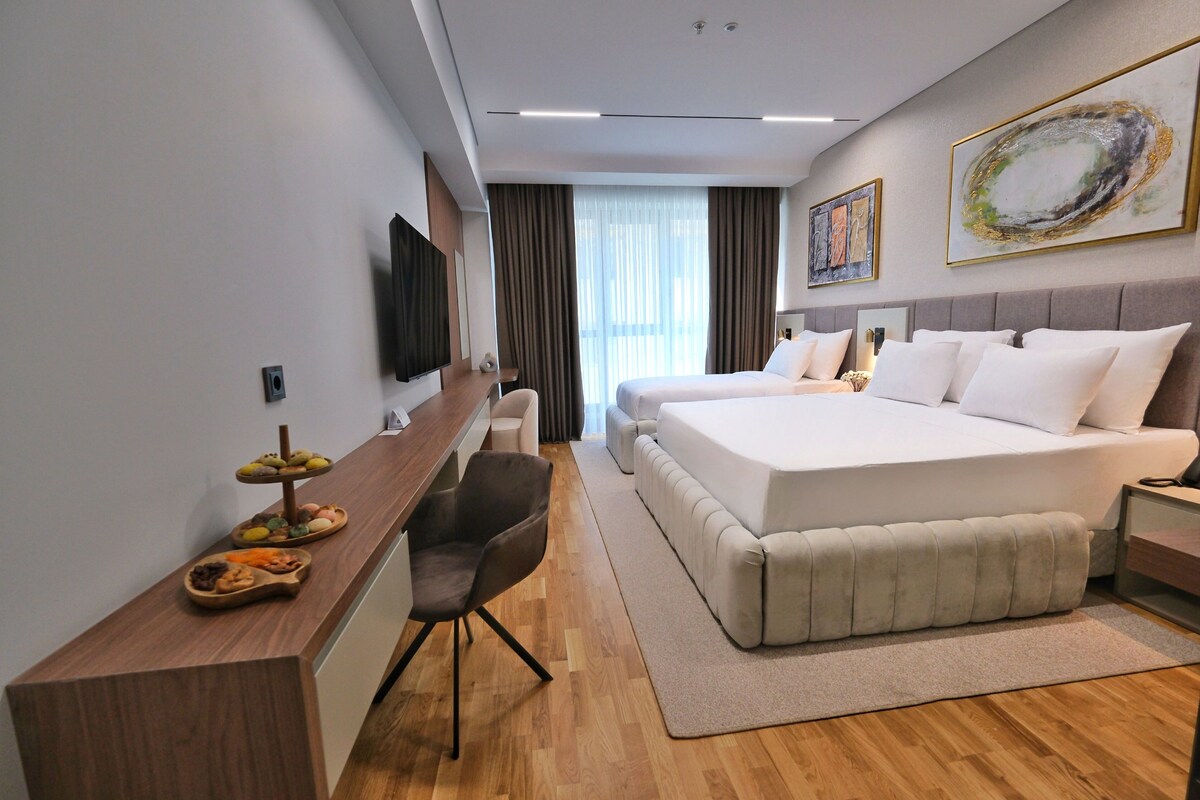 A modern hotel room with a large bed, a desk with a chair, a TV, and artwork on the walls.