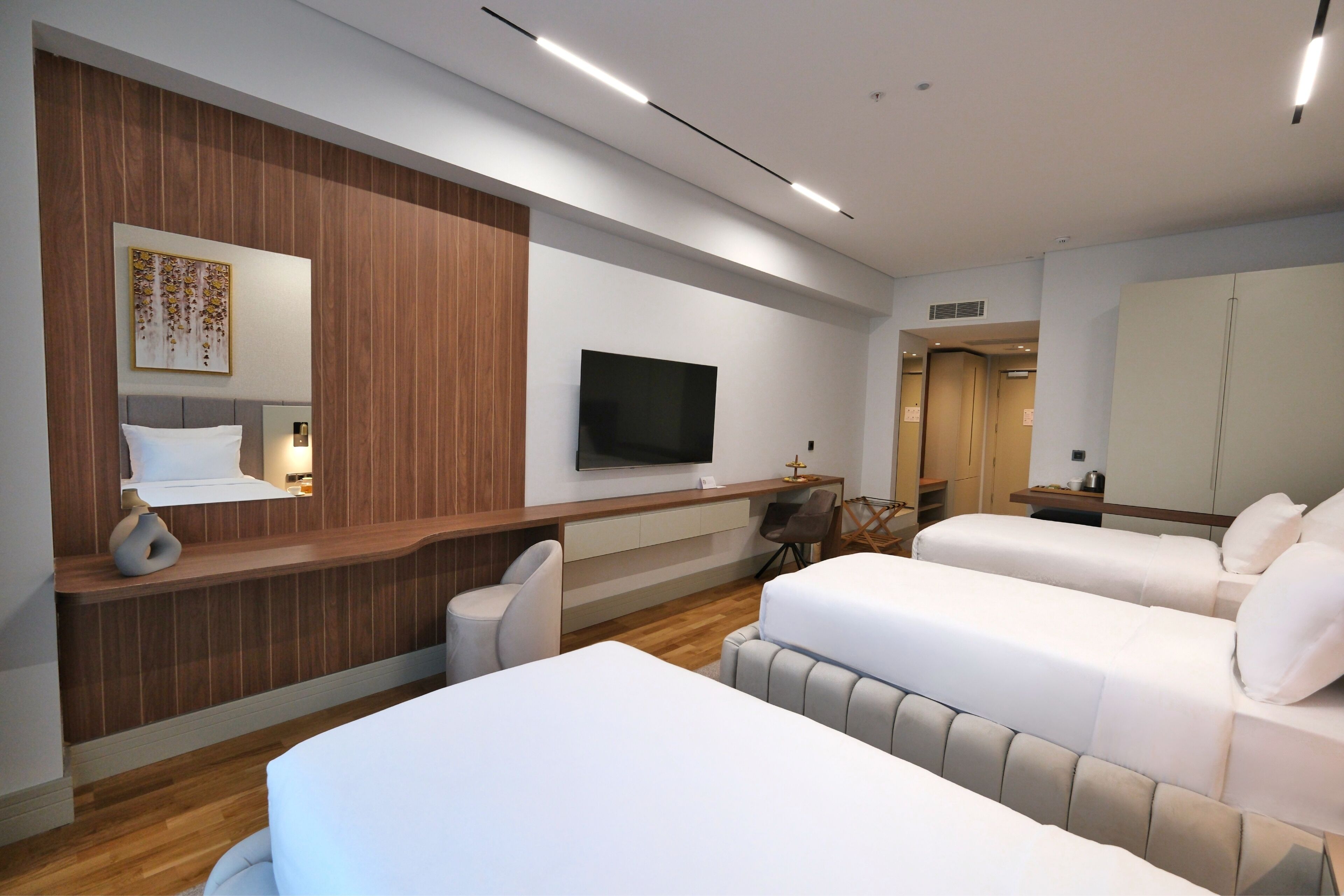 A modern hotel room with a wooden accent wall, a flat-screen TV, two beds, a desk with a chair, and a small table with a vase.