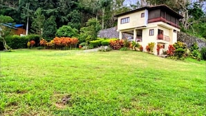 Una casa de dos pisos con balcón, rodeada de un césped cuidado y diversas plantas.