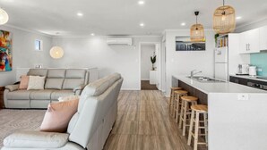 A modern living room with a grey sofa, a kitchen with a white countertop and bar stools, and a dining area with a table and chairs. - Wave Riders in Goolwa Beach (Goolwa Beach)