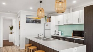 A modern kitchen with white cabinetry, a large island with stools, a built-in oven, and a hanging light fixture. - Wave Riders in Goolwa Beach (Goolwa Beach)