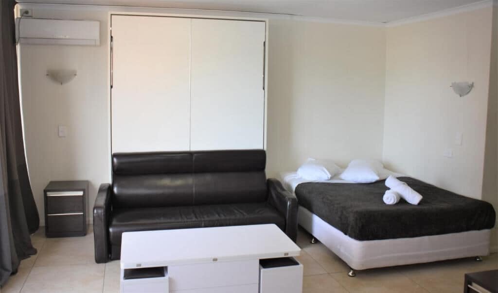 A compact room with a black leather sofa, a bed with a dark blanket, a white bedside table, and a small white cabinet.